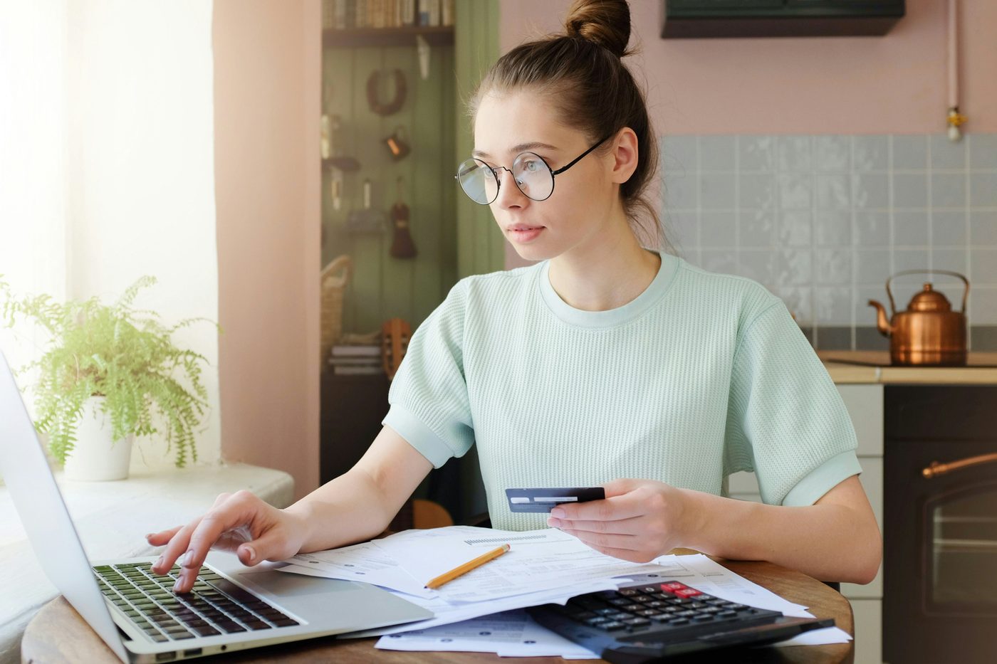 A woman at a kitchen table entering figures on a laptop with a card and calculator beside her