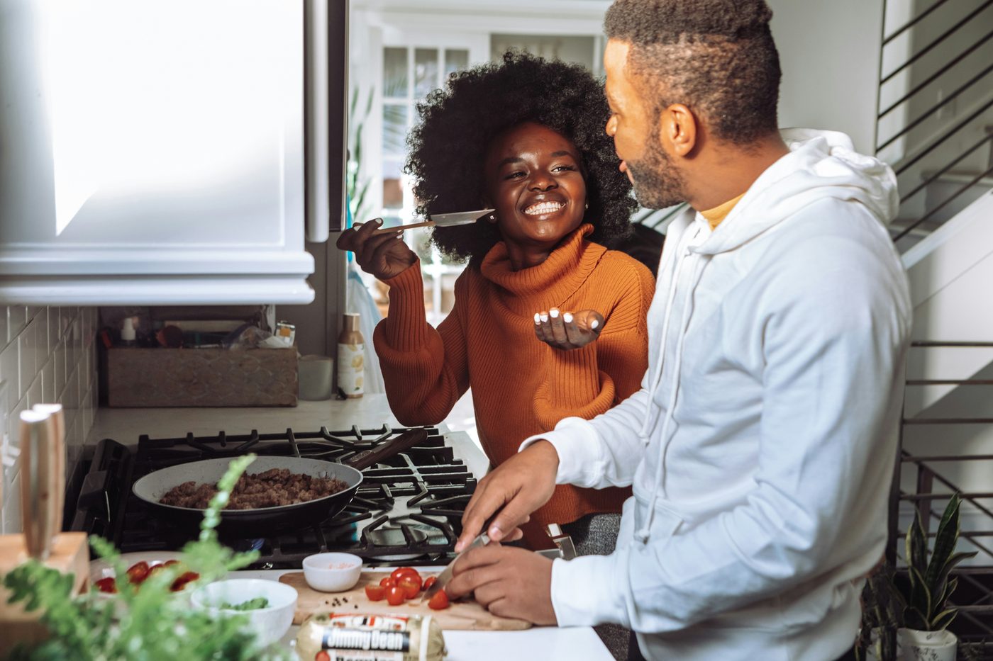 A couple cooking together at the stove, tasting from a spoon